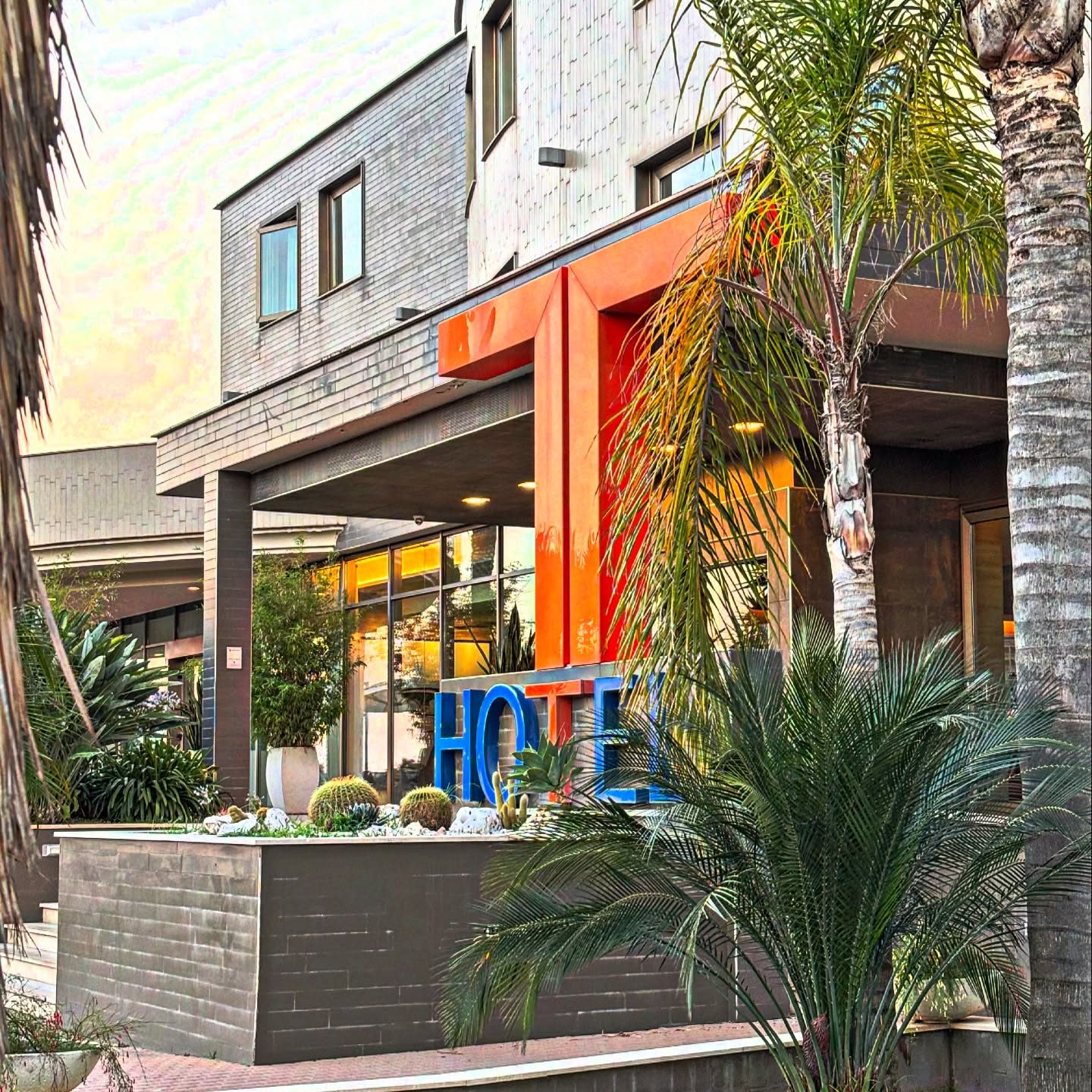 Ingresso del T-Hotel con grande T arancione, scritta HOTEL blu, palme e cactus su fioriera in pietra grigia
